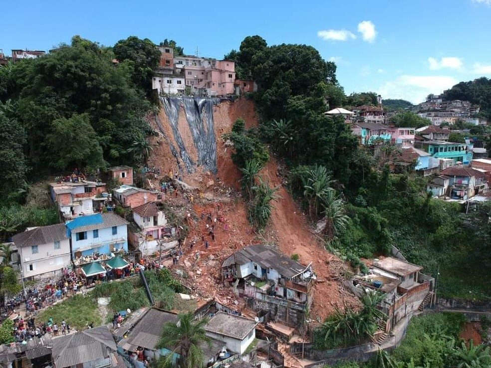 Vista aérea de um deslizamento de terra que destruiu diversas casas numa comunidade construída em encosta, com uma extensa cicatriz de terra exposta descendo o morro entre construções, e equipes de resgate e moradores reunidos na base do deslizamento.