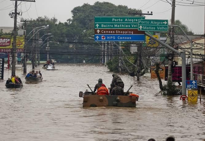 Militares em veículo blindado e moradores em barcos navegam por rua completamente alagada no bairro Mathias Velho, em Canoas (RS), durante as enchentes de maio de 2024; placas de trânsito indicam "Bairro Mathias Velho", "Canoas Shopping" e "HPS Canoas".