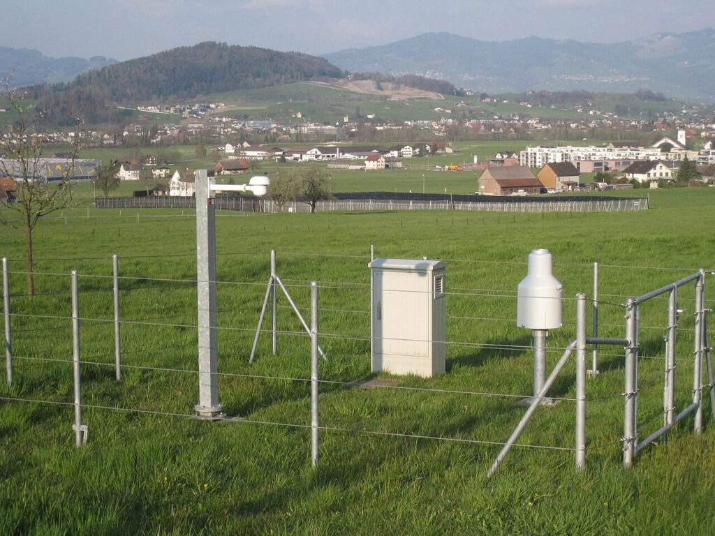 Estação meteorológica automatizada em campo aberto, na Suíça, com equipamentos de coleta de dados incluindo um pluviômetro — imagem ilustrativa do tipo de infraestrutura usada em redes de monitoramento de chuva, não é do caso específico nem do Brasil.